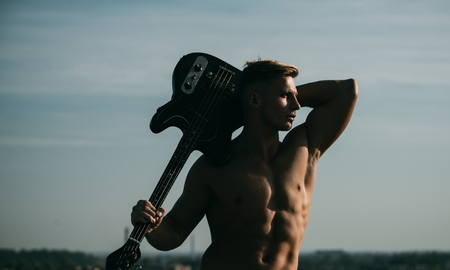 Man with muscular torso hold electric guitar on blue sky. Athlete guitarist with six pack, ab outdoor. Music, performance, entertainment. Sport, fitness, health concept. Training and playing.の写真素材