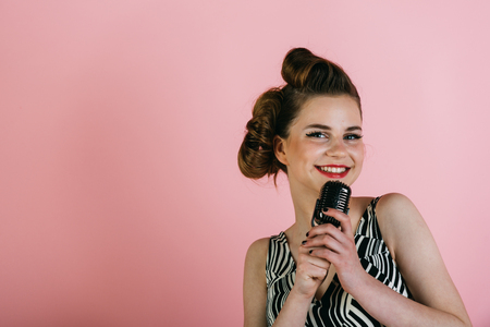 Girl in glasses sing in microphone. Woman singer with stylish retro hair and makeup. Music, look and retro style, pinup. Beauty and vintage fashion. Pin up young girl on pink background, radio.の写真素材