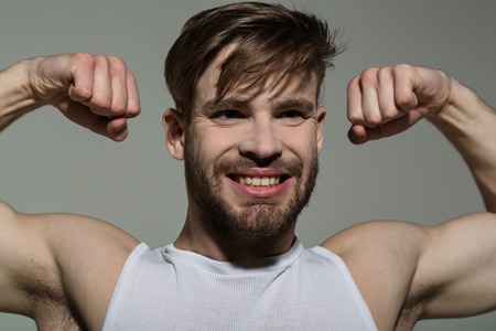 Macho smile with strong arms in white singlet. Man flex muscles, biceps, triceps on grey background. Sport, fitness, health. Workout, training, gym. Power, strength concept.の写真素材