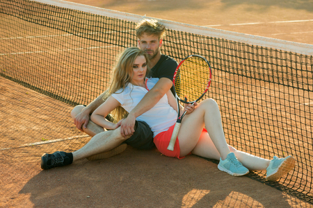 Activity, energy, health. Couple in love sit at tennis net on clay court. Relationship, relations, family. Sport, game, match concept. Woman and man tennis players with racket.の写真素材