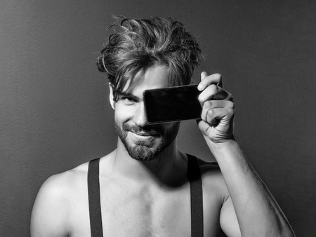 Smiling handsome man fashion sexy young blond bearded male model with suspenders on topless sexi nude torso hides face behind smartphone or mobile phone on grey backgroundの写真素材