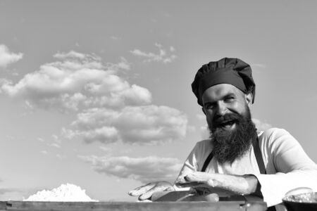 Man cook chef with beard on handsome smiling face in white and red uniform cooking dough sunny...