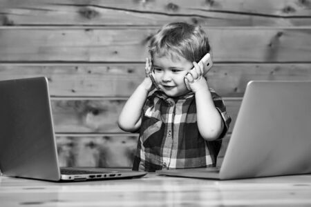 Cute smiling baby boy child with blond curly hair talks on two phones and plays on laptop computers on wooden backgroundの写真素材