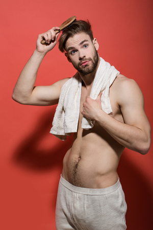 Guy brush hair with hairbrush on red background. Man with surprised face, bath towel in sexy underwear. Haircare, wellness, health. Beauty, grooming, hygiene. Morning routine concept.の写真素材