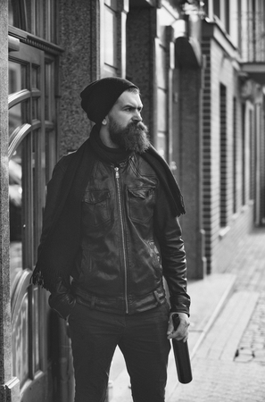 bearded man, long beard, brutal caucasian hipster with moustache holding glass wine bottle, has serious face, in black leather jacket, hat, scarf, unshaven guy with stylish hair near building outdoorの写真素材