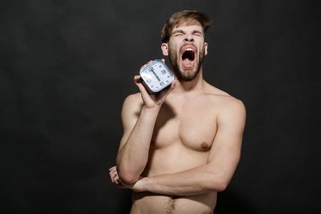 Sleepy macho with muscular torso, biceps, triceps. Man with alarm clock yawn on black background. Time, routine, deadline. Morning, wake up concept. Health, fitness, bodycare.の写真素材