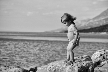 Cute baby boy child with curly blond hair in green shirt plays at rocky sea beach on summer dayの写真素材