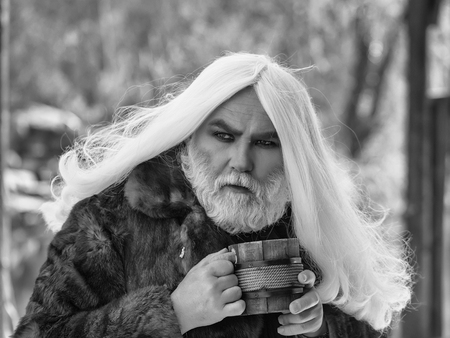 Druid old man with long grey hair and beard in fur coat with wooden mug in hands on blurred backgroundの写真素材