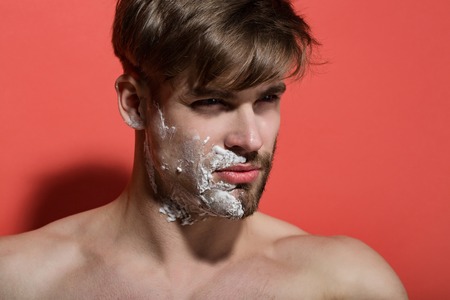 Man face half shaved and bearded with shaving cream on red background. Morning hygiene concept. Skincare, beauty, grooming.の写真素材