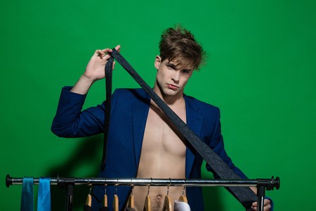 Businessman with tie in blue jacket on bare torso. Man choose necktie on wardrobe rack on green background. Business fashion, style concept. Clothing, dressing, closet. Shopping, sale, purchaseの写真素材