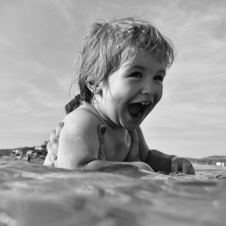 Cute laughing little boy bathes in clear water of sea on blue sky background.の写真素材