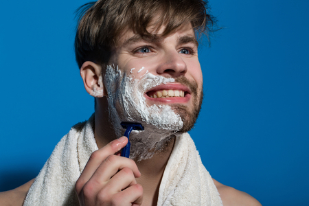 Bachelor smile with shaving cream, razor and towel. Happy man shave bearded face on blue background. Beauty, grooming, hygiene. Skincare, health, wellness. Morning routine concept.の写真素材