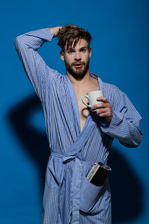 Guy with tea or coffee cup and magazine in dressing gown on blue background. Morning, breakfast conceptの写真素材