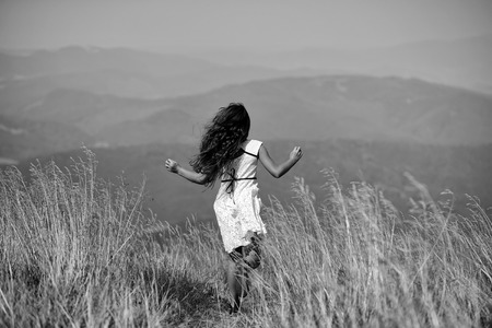 Back viev of young little brunette girl in white lace summer dress running in mountain valley with deep dry spikelet grass in spring sunny day outdoor on natural blue background, horizontal pictureの写真素材