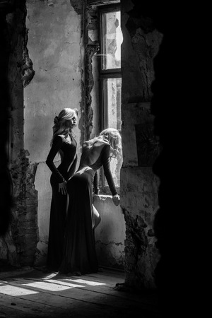young pretty women with long lush curly blonde hair and thoughtful face in green dress standing near stony wall and big window sunny dayの写真素材