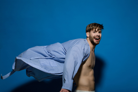 Happy bachelor with sexy torso in flying bathrobe. Man smile in dressing gown on blue background. Home clothes, leisure wear. Spa, wellness, grooming. Fashion, style concept.の写真素材