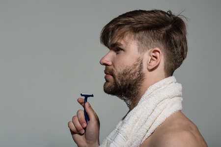 Guy with bearded face profile with razor and towel on grey background. Morning routine concept. Beauty, barber, hairdresser salon. Grooming, hygiene, skin care, copy spaceの写真素材