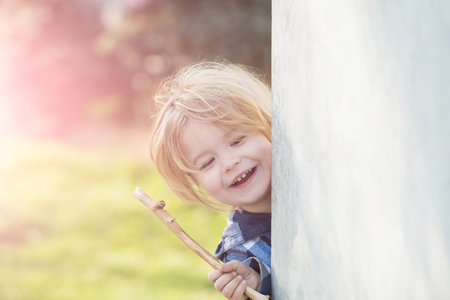 Kid with blond hair smile with stick at wall corner on sunny day on natural background. Happy child, childhood, playtime, lifestyle conceptの写真素材