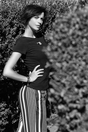 Pretty girl young beautiful woman with brunette hair in black tshirt and striped trousers poses outdoors in green bushes on sunny summer dayの写真素材