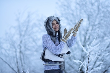 Woman skater with figure skates in fur hat, mittens, sweater smile in snowy forest outdoor. Winter fashion, style. Vacation, holidays, hobby, lifestyle. Ice skating, sport, activity, health concept.の写真素材