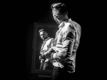 handsome muscular macho man or athlete guy with sexy body of bodybuilder has beard on serious face in pants, white shirt near vintage mirror isolated on black backgroundの写真素材