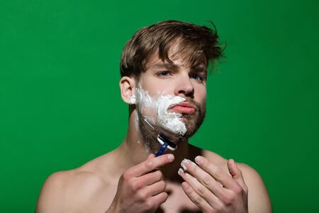 Skincare, health, wellness. Bachelor with shaving cream or foam on face skin. Beauty, grooming, hygiene. Morning routine concept. Man shave beard hair with razor on green background.の写真素材