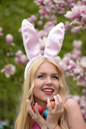 easter eggs in had of happy girl or cute woman with long, blond hair, in rosy, bunny ears on blossoming magnolia, floral environment. Spring. Easter. Holidays, celebrationの写真素材