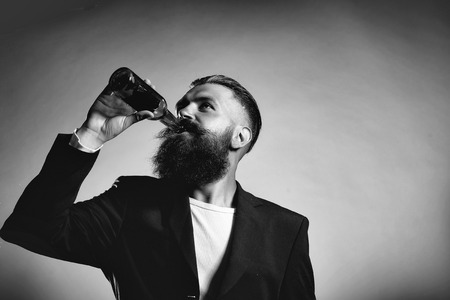 Handsome young man with long beard and moustache in black jacket holding green glass beer bottle in studio on grey backgroundの写真素材