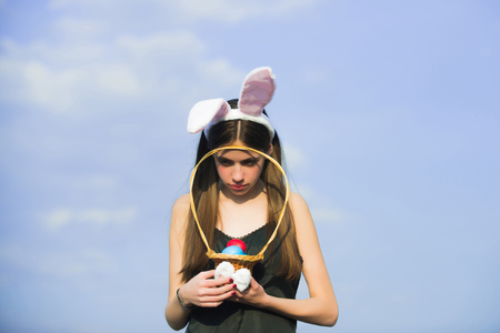 Easter woman holding wicker basket with colored eggs. Unhappy girl with rosy bunny ears on blue sky....