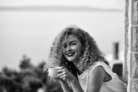 pretty woman with curly blond hair stands on balcony with coffee cup summer day on natural backgroundの写真素材