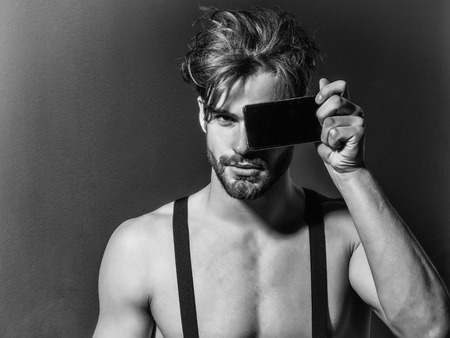 Handsome man confident fashion sexy young blond bearded male model with suspenders on topless sexi nude torso hides face behind smartphone or mobile phone on grey backgroundの写真素材