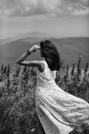 Pretty cute sexy brunette woman or girl stands back in blue dress waving on wind touches her hair outdoor on beautiful landscape on blue sky backgroundの写真素材