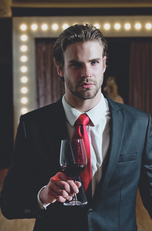 businessman or man with beard and hairstyle in formal outfit with red tie hold wine glass near mirror with lamps, meeting and relax, bar and restaurant, sommelier, tasting and degustationの写真素材