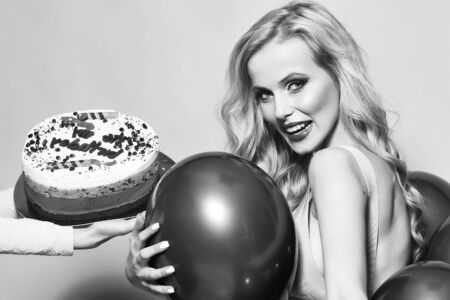 One attractive smiling young happy blond woman with long curly hair with birthday cake with candle in female hand near bunch of red party balloons in studio on yellow backdrop, horizontal pictureの写真素材