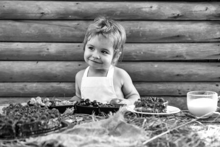 Cute little boy eats green apple at table with berries raspberry gooseberry cherry currant fruit cake and cup of milk on wooden backgroundの写真素材