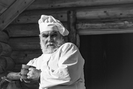 bearded man cook chef in uniform and hat with long beard holding wood cup with iron decoration sunny day outdoor on wooden backgroundの写真素材