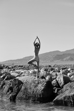 Young straight healthy woman in summer cloth standing in pose of yoga outdoor sunny day on stones near water coast on natural background, vertical pictureの写真素材