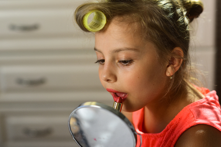 Beauty and fashion concept. Child with nice cheeks smiles and paints lips indoors. Little girl holds lipstick and mirror sitting in room. Kid tries to paint lips and do makeup with mothers cosmetics.の写真素材