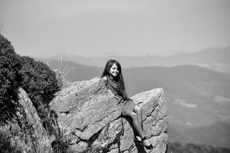 Beautiful brunette smiling girl touching long hair in blue lace dress sitting on stone rock cliff looking at beautiful landscape sunny day outdoor on natural blue sky background, horizontalの写真素材