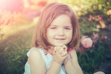 Germination and growth. Innocence, purity and youth concept. Child sitting at blossoming rose flowers on green grass. Girl smiling with folded hands in summer garden. Future and flourishing.の写真素材