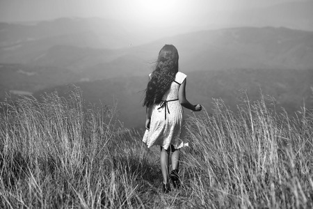 Back viev of young small brunette girl in white lace summer dress walking in mountain valley with deep dry spikelet grass in spring sunny day outdoor on natural blue background, horizontal pictureの写真素材