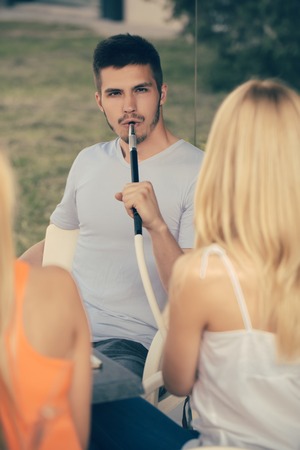 Man vapor hookah pipe with girls in shisha bar lounge. Celebration, party concept. Addiction, bad habits. Date, love triangle, relations.の写真素材
