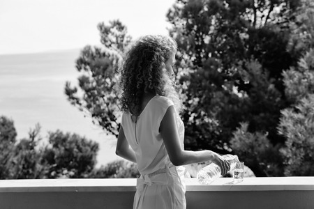Pretty woman or girl with curly blond hair pours pure drinking water from plastic bottle into glass on balcony on summer day on natural backgroundの写真素材