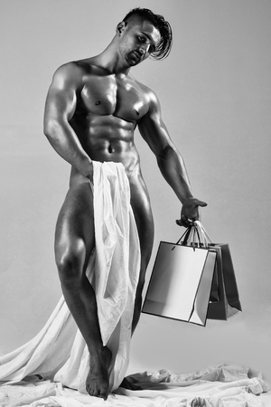 handsome muscular man or naked macho guy with sexy wet athletic body and torso, hides under white textile cloth or fabric, holds gift shopping bags on grey backgroundの写真素材