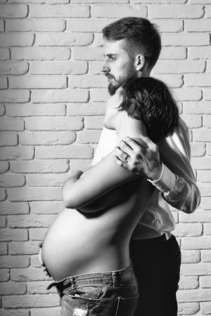 family parent couple of pretty smiling sexy woman or cute pregnant girl with naked round belly or abdomen and handsome bearded man hipster in white shirt on brick wall background at mothers dayの写真素材