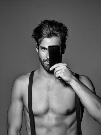 Handsome man confident fashion sexy young blond bearded male model with suspenders on topless sexi nude torso hides face behind smartphone or mobile phone on grey backgroundの写真素材