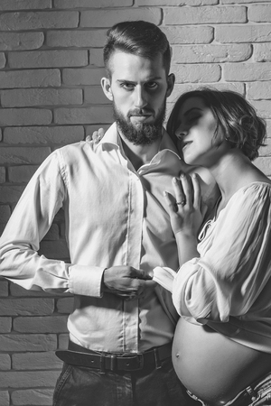 family parent couple of pretty happy sexy woman or cute pregnant girl with round belly or abdomen and handsome bearded man hipster in white shirt on brick wall background at mothers dayの写真素材
