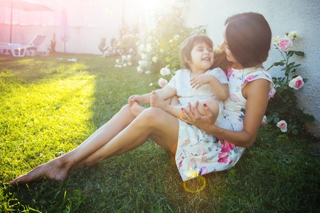 Nurturing and future. Mothers day concept. Woman with baby girl sitting on green grass. Family love and care. Mom and child smiling at blossoming rose flowers.の写真素材