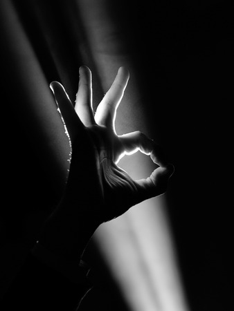 dark silhouette of human male hand with fingers in spotlight or backlight light with okay or ok gesture on black background with dramatic projector shine ray or beamの写真素材
