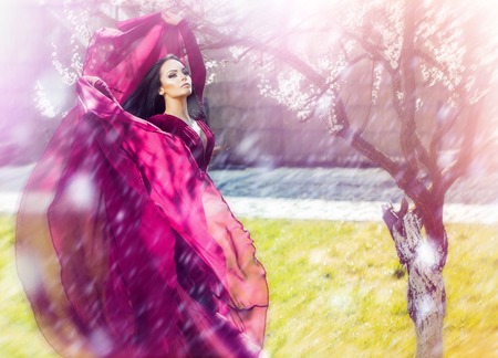 Sensual woman in violet dress in spring garden, soft filter. Pretty girl with flower petals falling from tree. Fashion, style concept. Beauty, look, makeup, long hair. Spring, nature, youth.の写真素材
